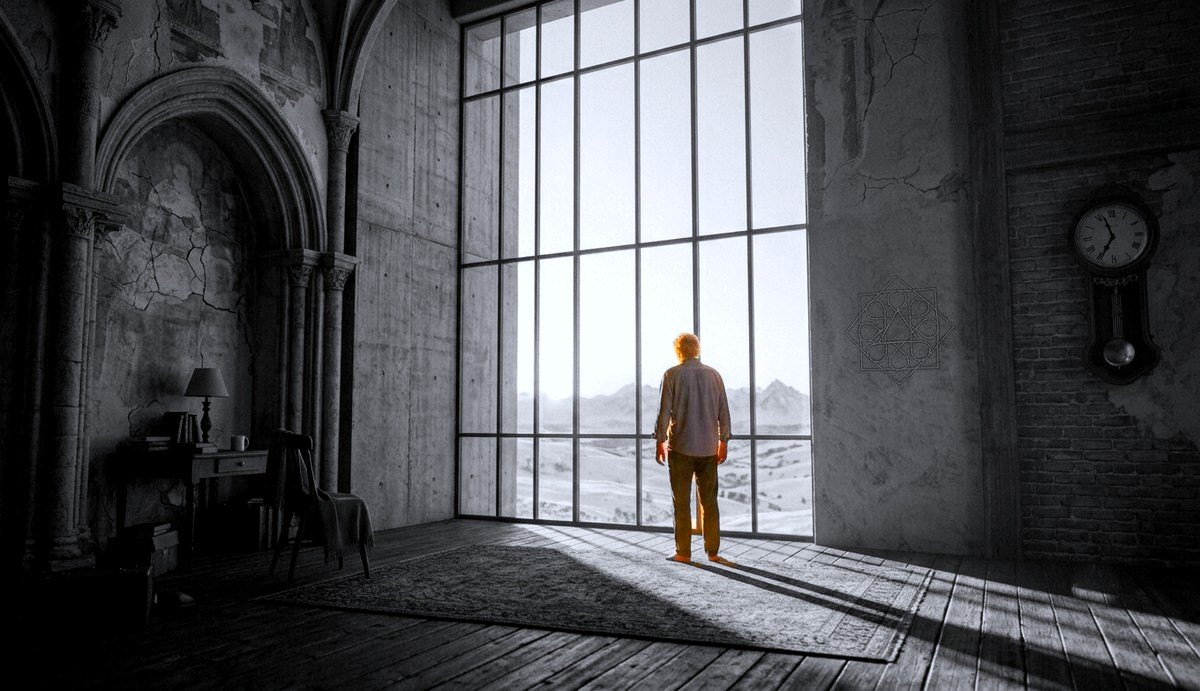 A man stands in a large grey room looking out of a tall window at a low sun